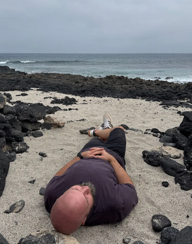 Tom lying back on a sandy beach among dark rocks, with the sea behind him.